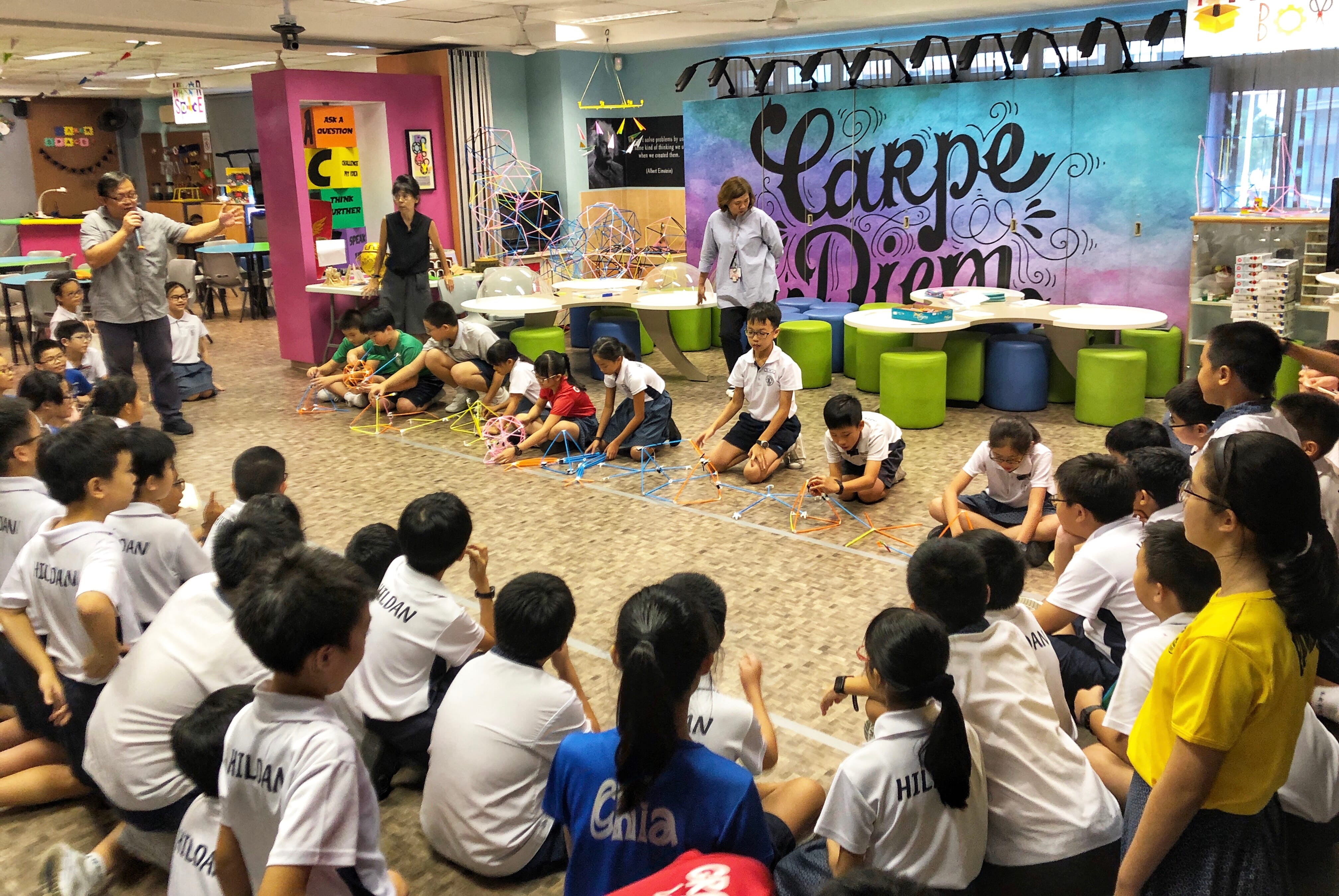 Students sit in a circle as classmates build straw structures on the floor; Carpe Diem mural behind.